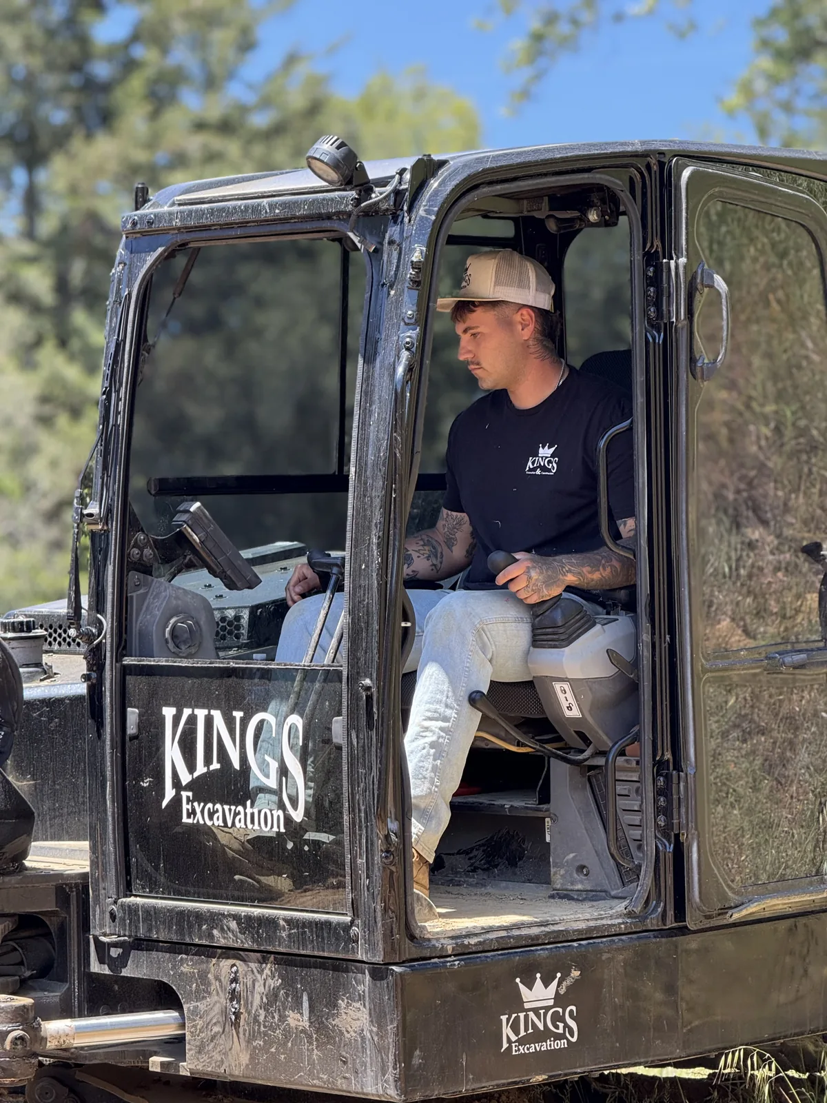 Kings Excavation team member in a Kings-branded excavator cab on a wooded jobsite